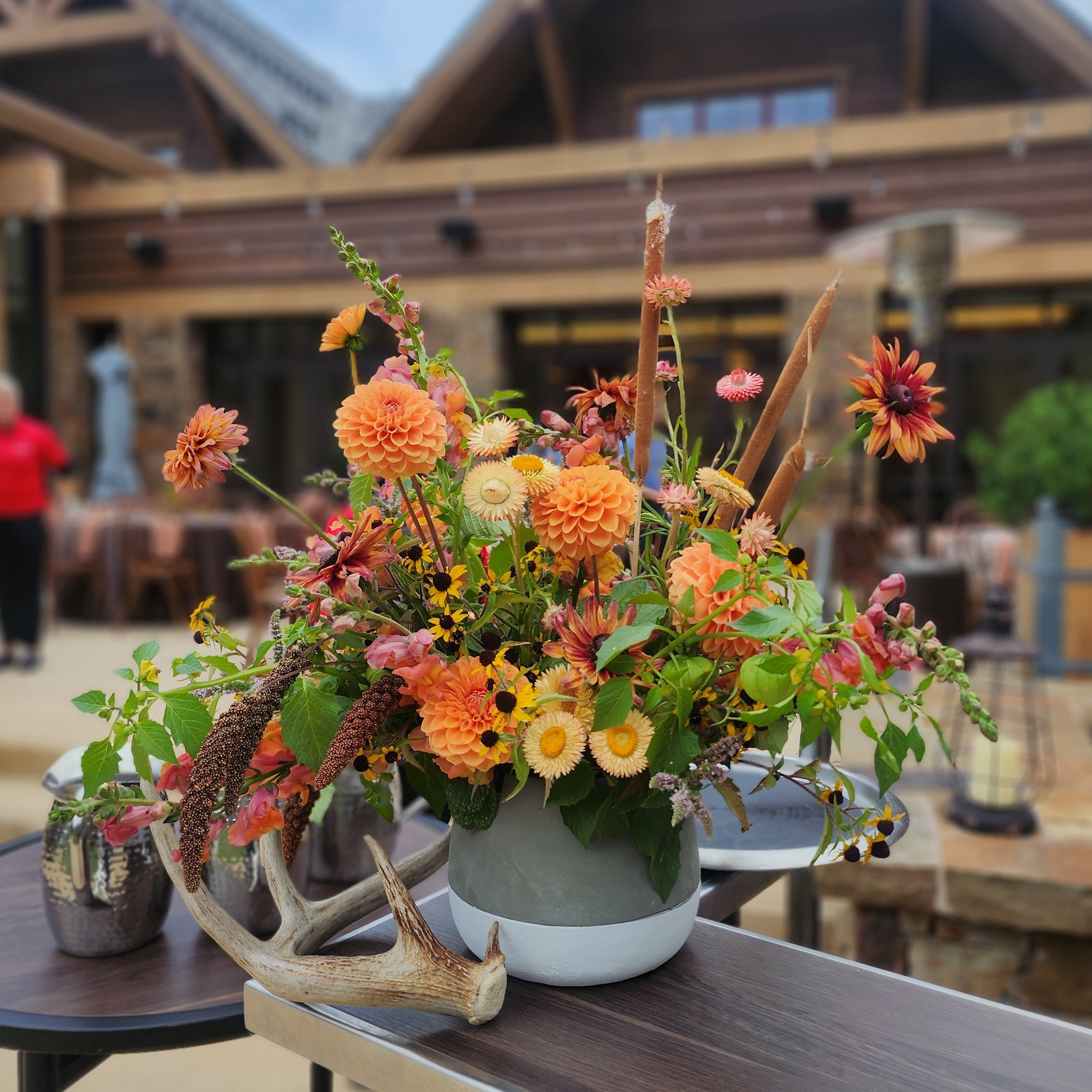An autumnal floral arrangement in a white pot with a deer antler, displayed on a wooden table in front of a rustic lodge building.